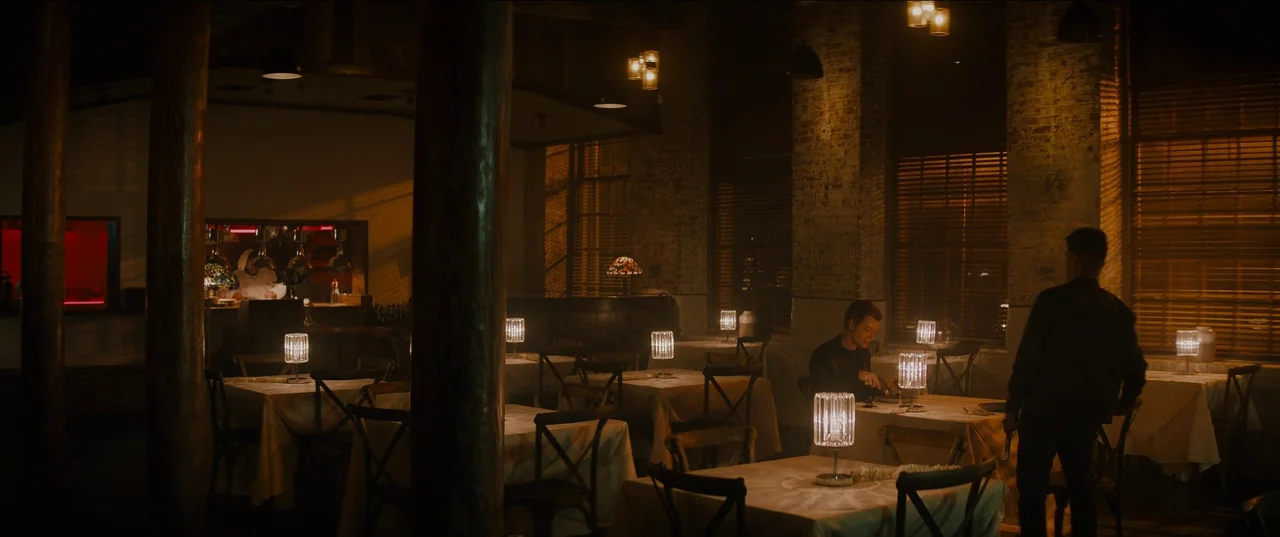 Night Teeth (2021) movie still: wide — A dimly lit upscale dining area with white-clothed tables and large wooden support pillars. A man eats alone…