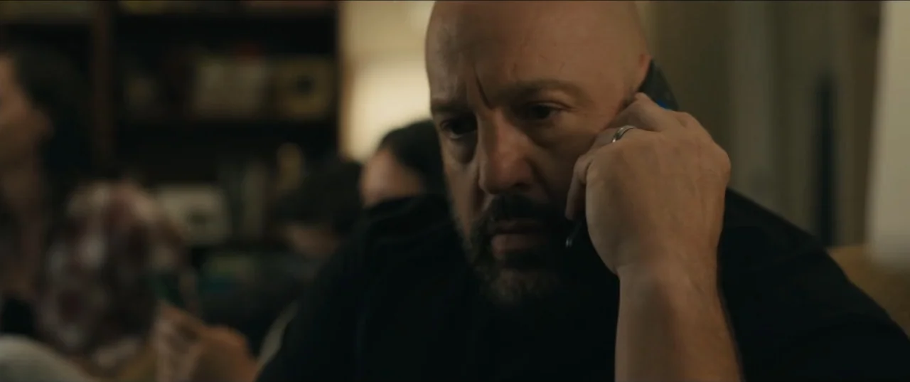 Guns Up (2025) movie still: closeup — An interior space with a blurred, dimly lit background suggesting a home environment. A bald man in a black…