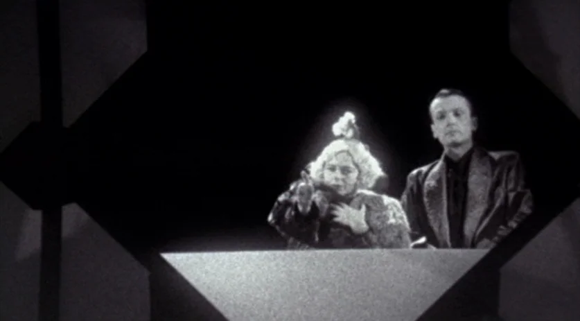 The Saddest Music in the World (2003) movie still: medium — A dark television studio set features two individuals positioned behind a geometric…