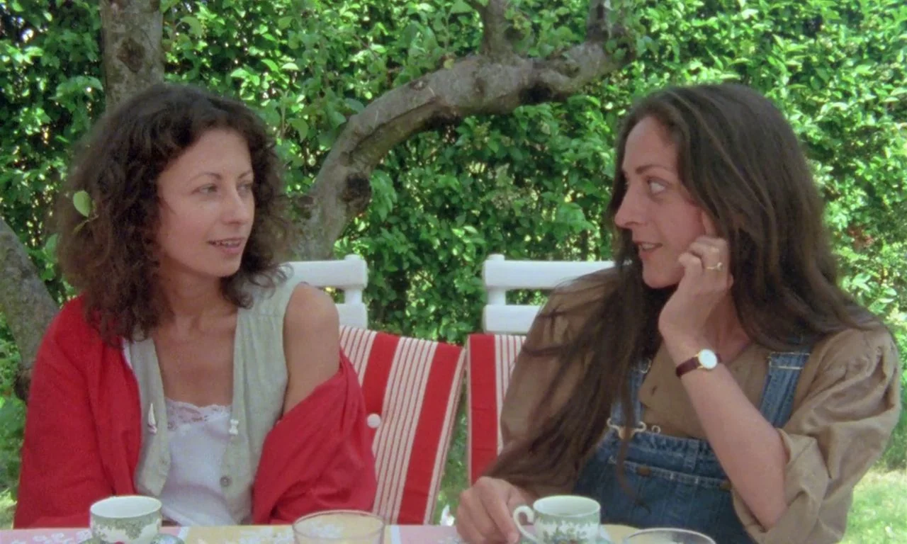 The Green Ray (1986) movie still: medium — Two women converse while sitting at a table beneath the canopy of a leafy tree. They are positioned…