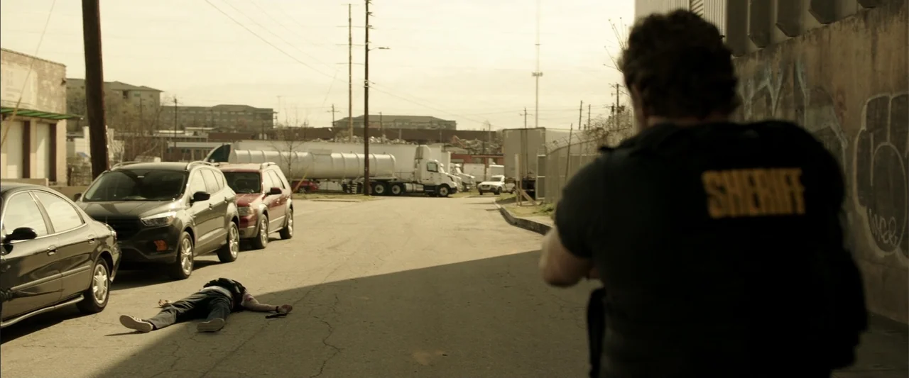 Den of Thieves (2018) movie still: medium — An industrial street with parked cars and a distant tanker truck. A sheriff stands in the foreground…