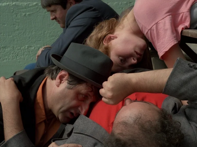 Keep Your Right Up (1987) movie still: medium — A group of people lies close together against a weathered green concrete wall. The individuals…