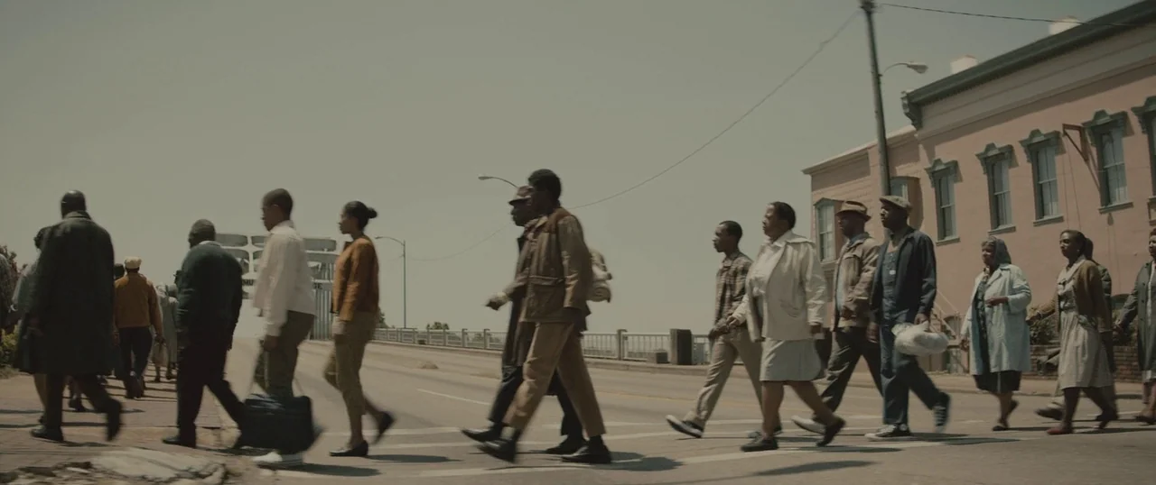Selma (2014) movie still: wide — A group of individuals in 1960s attire walks across an urban paved intersection. To the right stands a multi-story…