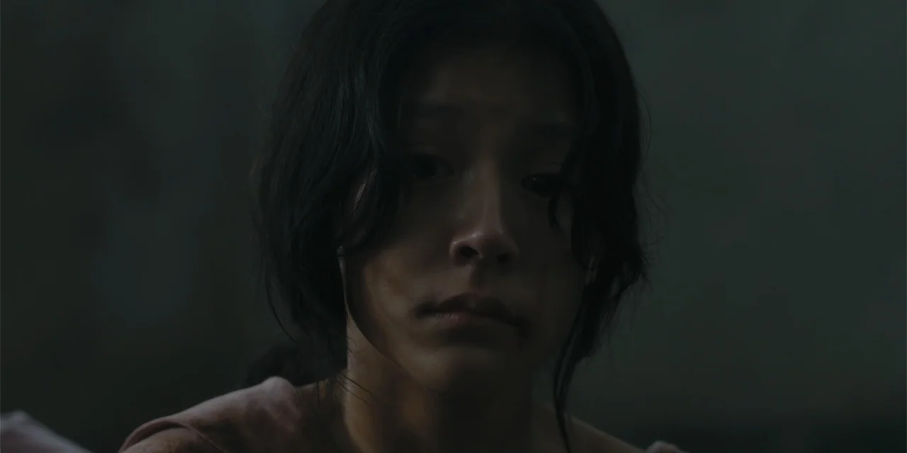 Host (2020) movie still: closeup — A dark and minimally lit interior space with shadows obscuring the surroundings. A young woman with dark hair…