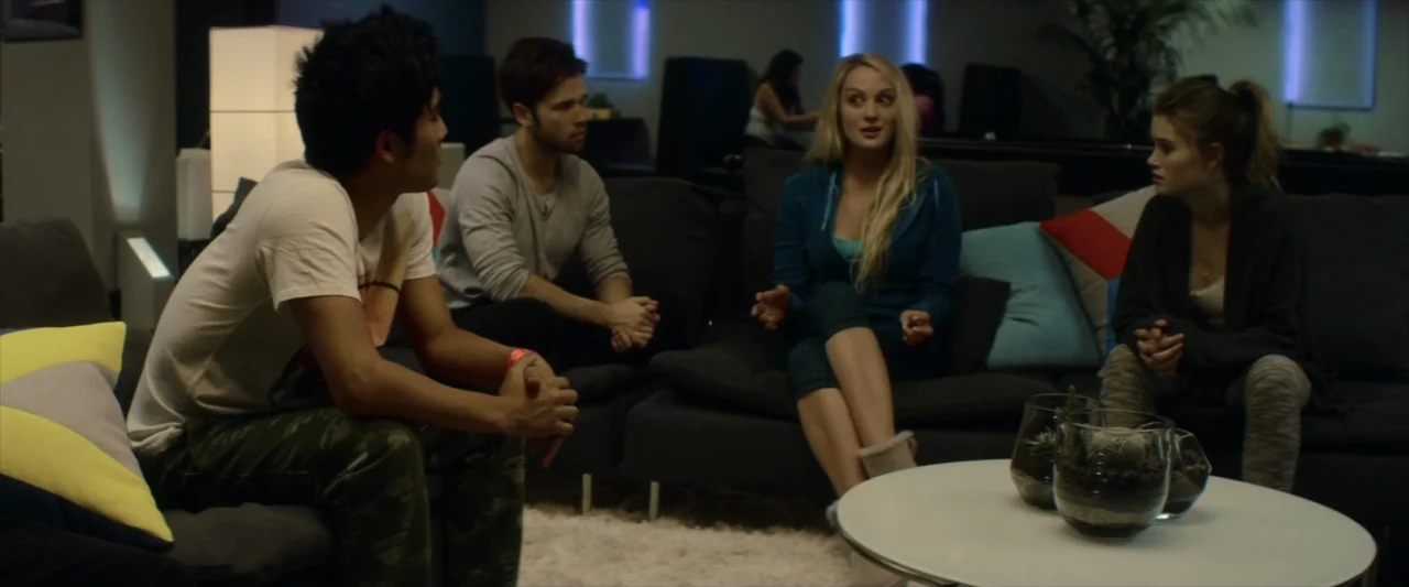 Tell Me How I Die (2016) movie still: medium — A modern lounge area features dark fabric sofas arranged around a circular white table. Four young…