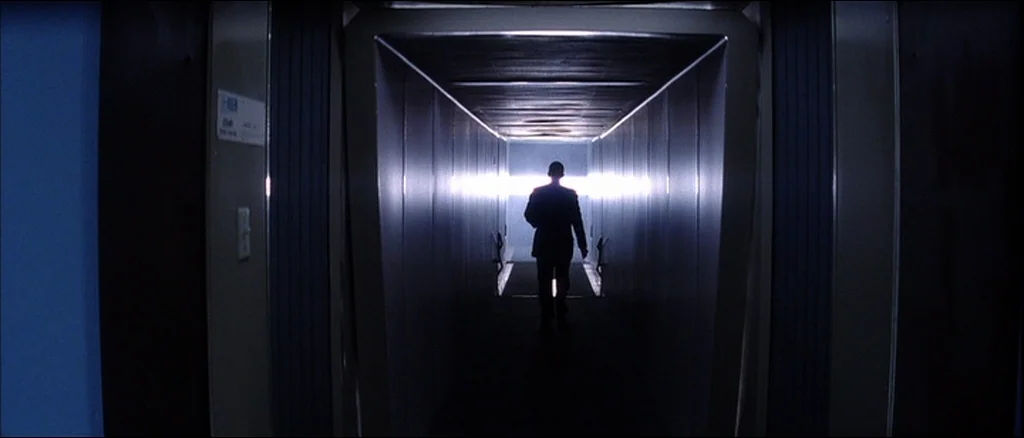 Punch Drunk Love (2002) movie still: fullbody — An enclosed jet bridge has metal walls and a narrow, rectangular passage. A solitary man in a dark…