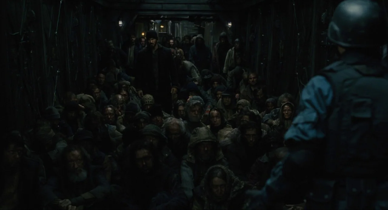 Snowpiercer (2013) movie still: wide — A long, dark metal interior of a transport vehicle filled with many seated people. A guard wearing tactical…