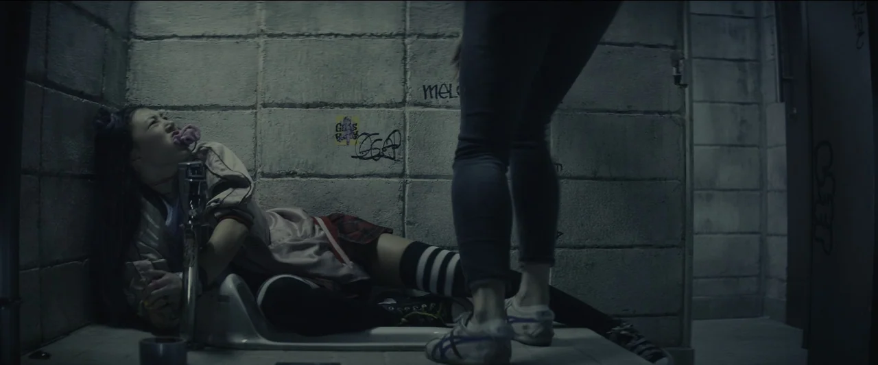 Kate (2021) movie still: medium — A public restroom stall features rough gray cinder block walls covered in dark graffiti tags. A young woman in…