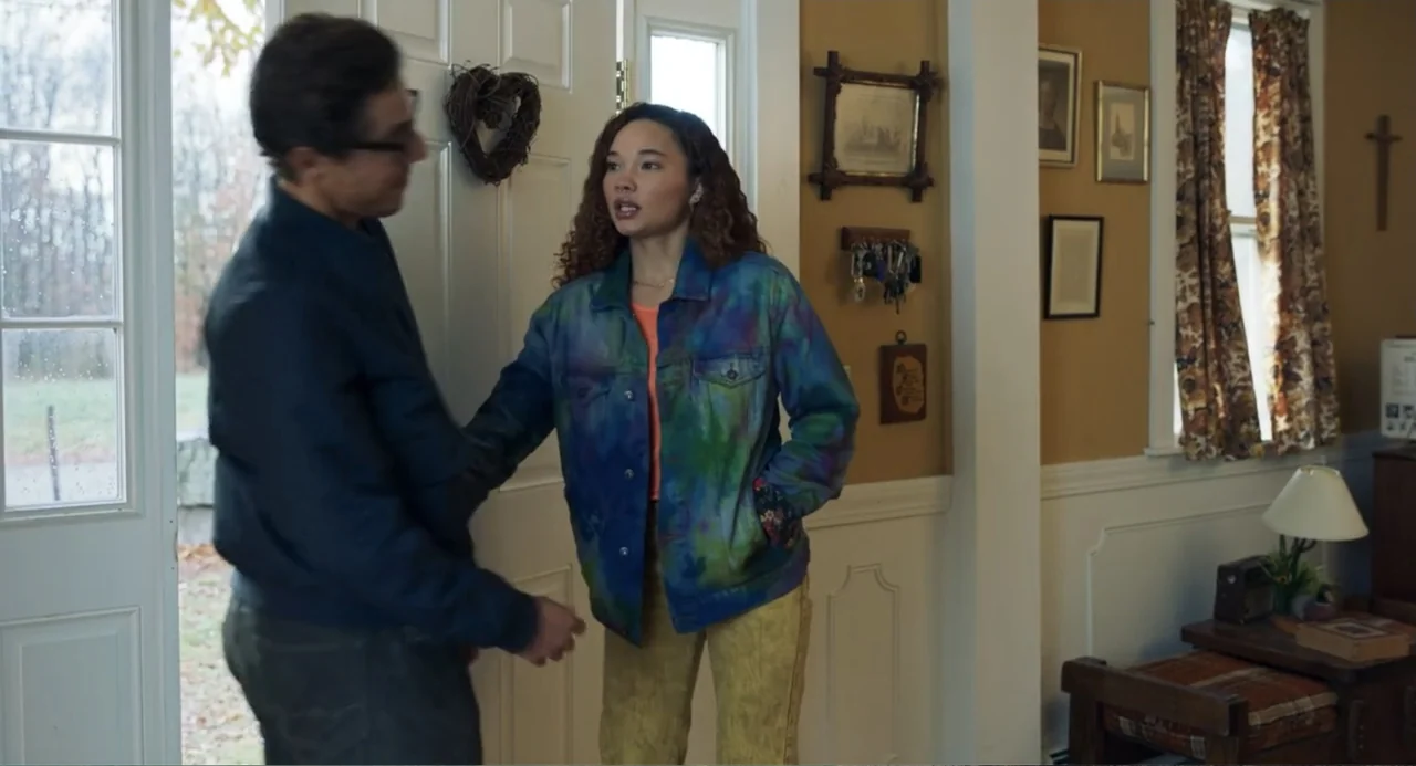 Salvation (2025) movie still: medium — A residential home entryway with tan walls features framed art and a key rack. A woman in a tie-dye jacket…