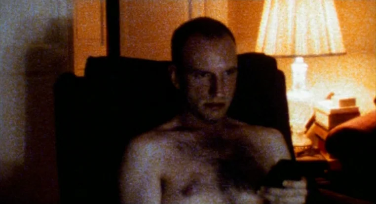 Schizopolis movie still: medium — A dimly lit room features a table lamp with a patterned shade next to a chair. A shirtless man sits in the dark…