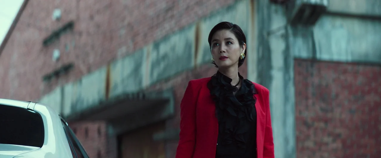 Believer (2018) movie still: medium — A woman stands before a weathered red brick industrial building with grey concrete elements. She wears a…