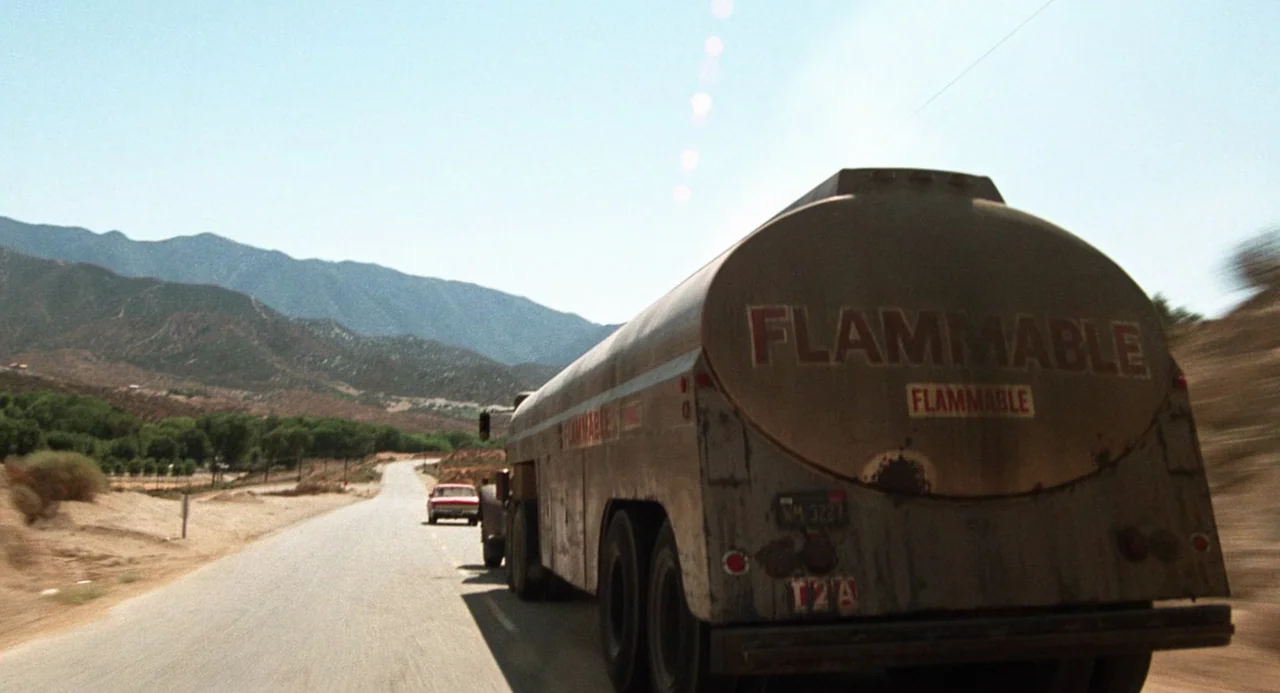 Duel (1971) movie still: wide — A weathered fuel tanker travels down a sun-drenched, paved desert road. Mountains rise in the background under a…