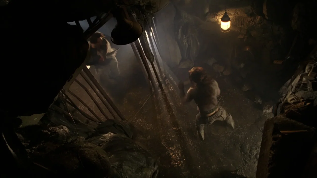 The Chronicles of Riddick (2004) movie still: medium — A dark, cramped dungeon space is defined by uneven stone walls and heavy vertical iron bars.…