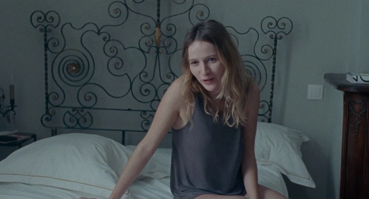 Non-Fiction (2018) movie still: medium — A quiet room contains a white bed with a large, ornate metal headboard. A young woman with long blonde hair…