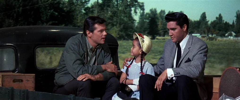 It Happened At The World’s Fair (1963) movie still: medium — Three individuals sit in the back of a truck traveling through a rural landscape. The…