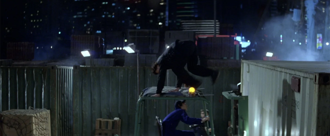 Rush Hour (1998) movie still: wide — A night time shipping yard with steel shipping containers and a forklift in motion. A person in black jumps…