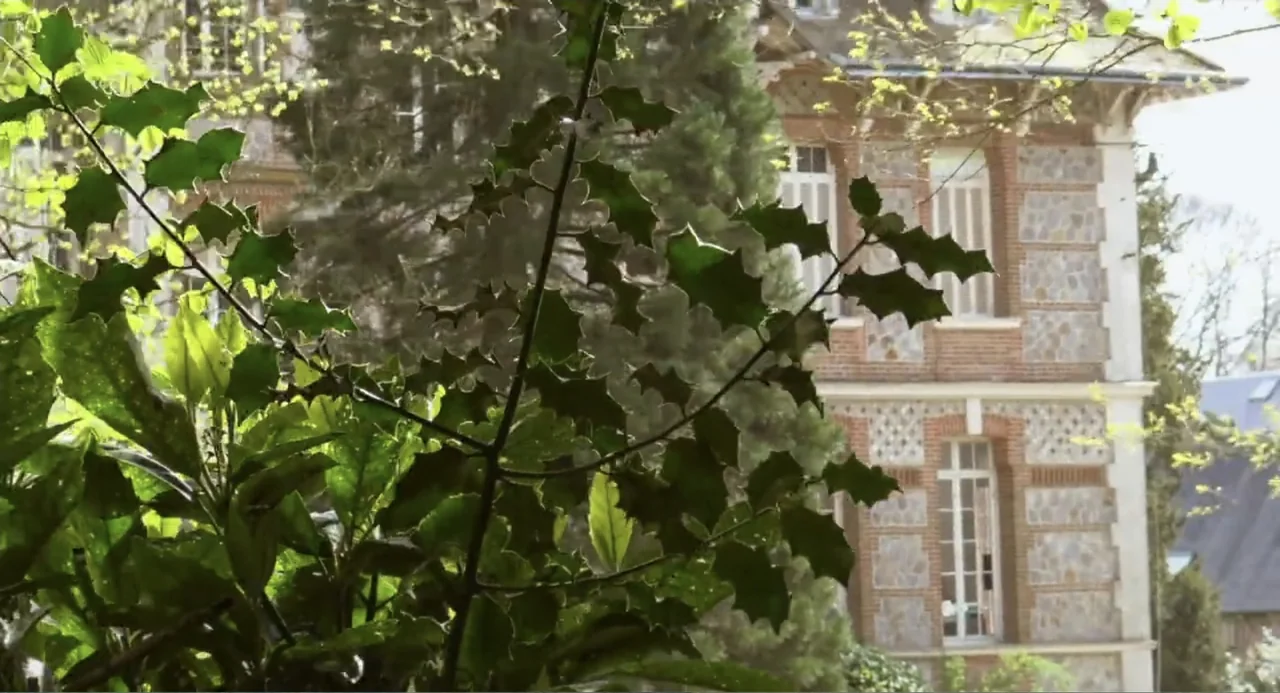 A Dramatic Film (2019) movie still: wide — A stone and brick house stands in the background behind dense green foliage. Lush bushes with holly…