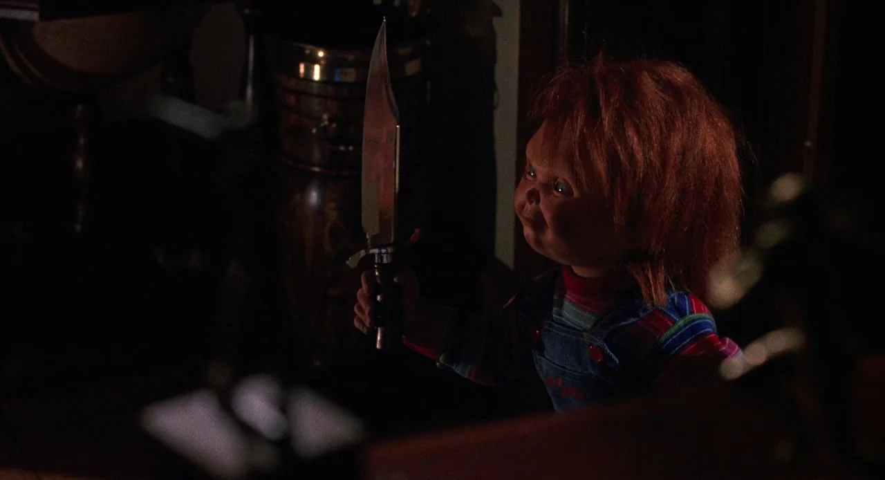 Child’s Play 3 (1991) movie still: medium — A dimly lit room holds a small plastic doll standing in the foreground shadows. The doll grips a large…