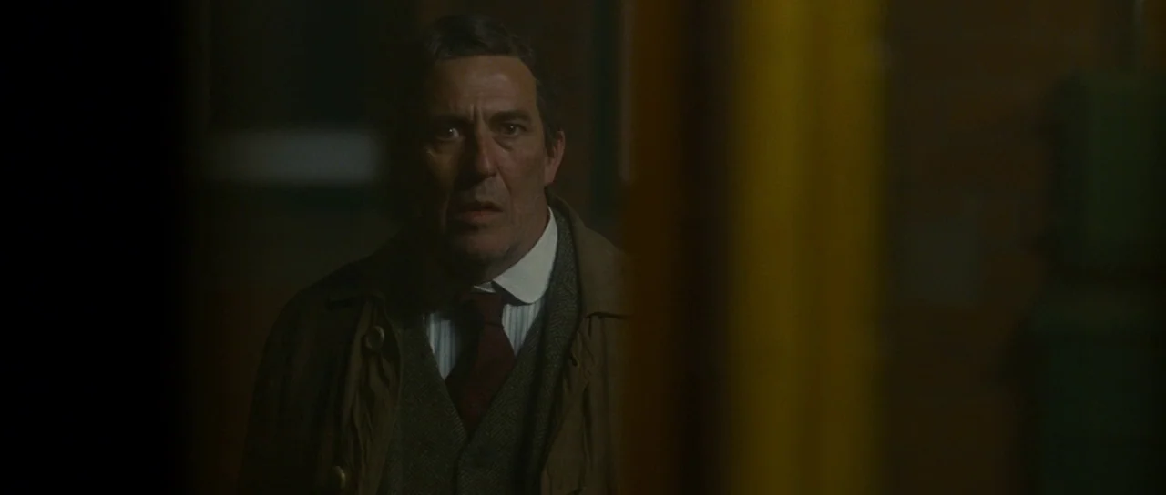 The Woman in Black (2012) movie still: medium — A dimly lit narrow hallway with a yellow partition partially obscuring the view. An older man in a…