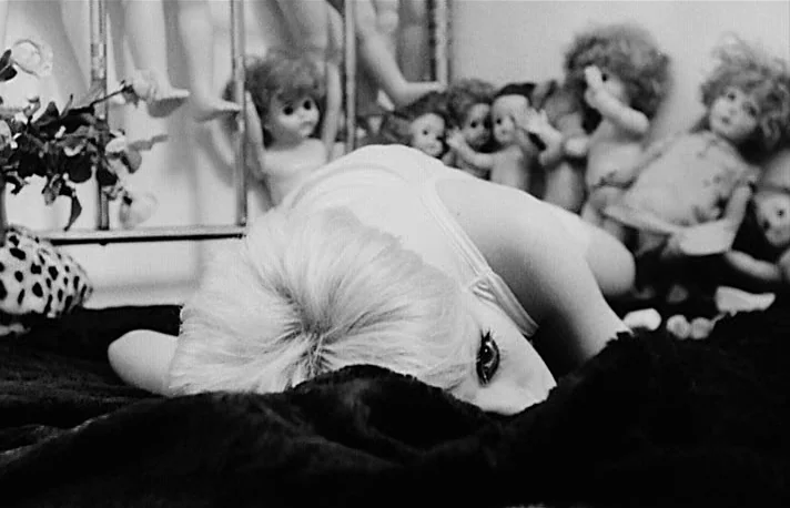 Fando y Lis (1968) movie still: medium — A woman lies prone on a dark fabric surface covered in numerous dolls. The dolls are arranged in the…