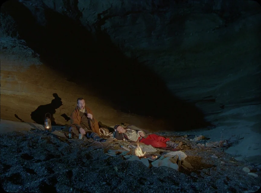 Jauja (2014) movie still: wide — A dark, rocky cave interior is illuminated by a small, low-burning campfire. Two men rest on the uneven dirt…
