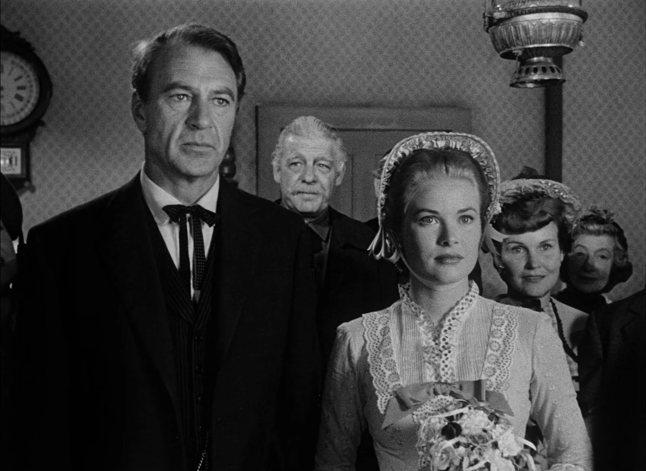 High Noon (1952) movie still: medium — An interior space with patterned wallpaper features a man and woman in formal attire. The couple stands…