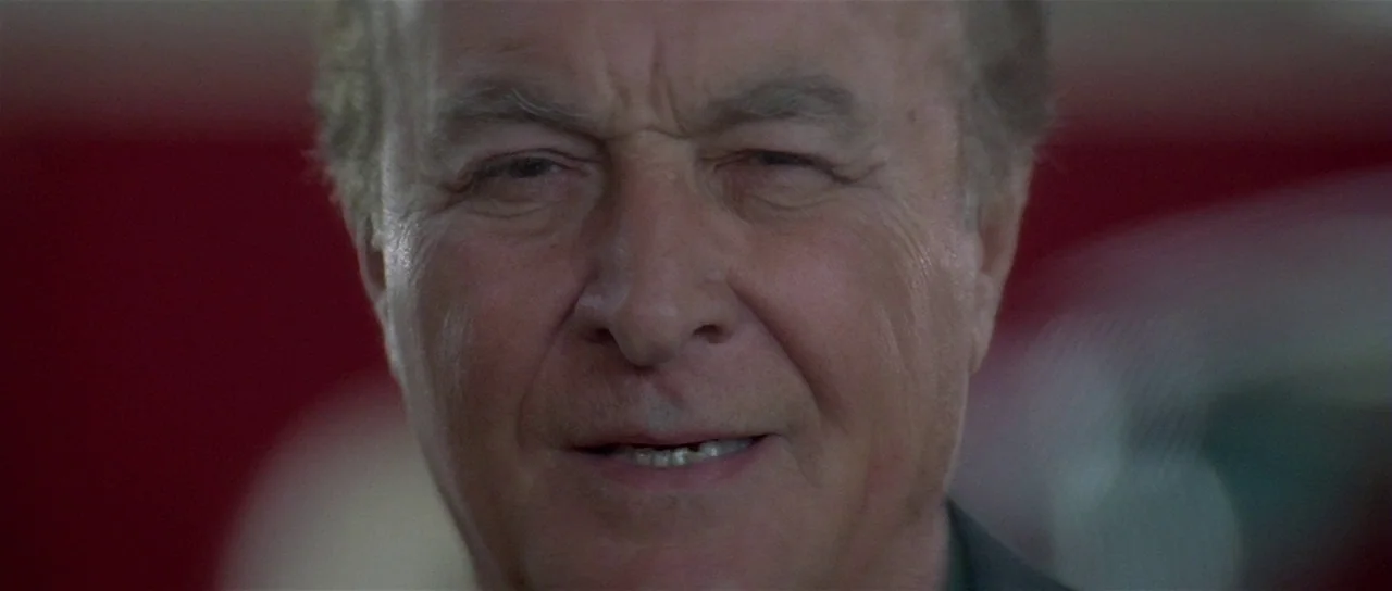 Lost Highway (1997) movie still: closeup — A man's face fills the frame against a blurred dark red background. The subject maintains eye contact…