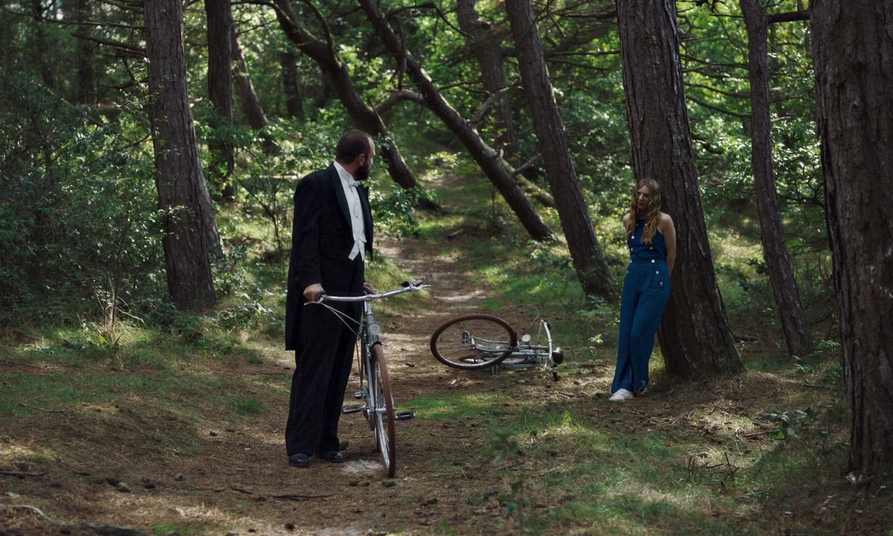 Bloodsuckers (2021) movie still: fullbody — A dense patch of pine trees with sandy ground covered in scattered pine needles. A man in formal wear…