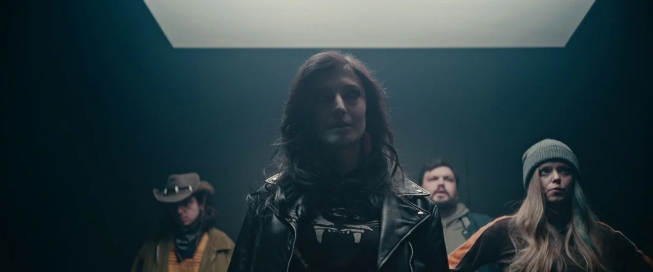 We Are Zombies (2023) movie still: medium — Four people stand inside a dark, confined space illuminated by an overhead light. They stare upward…