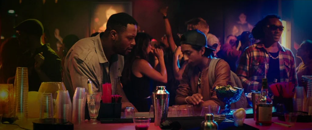 Dope (2015) movie still: medium — A dimly lit nightclub bar features colorful neon lighting and a crowded dance floor. Two men stand at the counter…