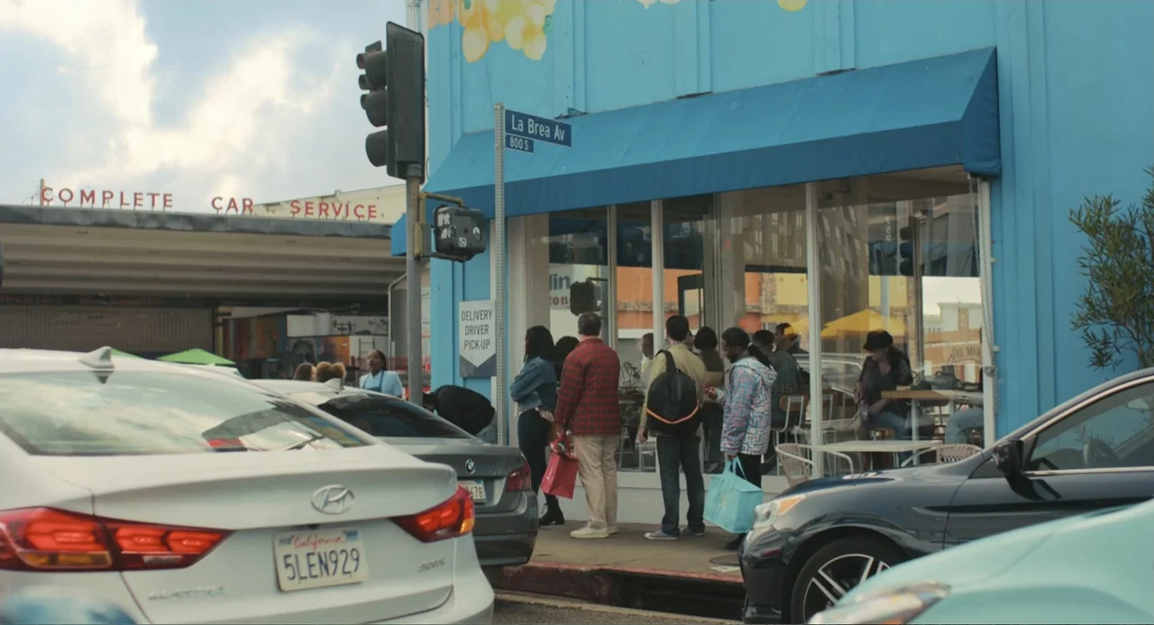 Good Fortune (2025) movie still: wide — A bright blue building exterior features a street corner restaurant entrance on a city block. A small group…