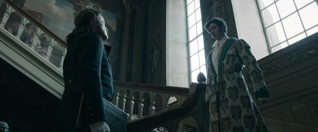 Napoleon (2023) movie still: fullbody — A grand interior staircase features ornate carved railings and a large mural on the wall. Two men, one in a…