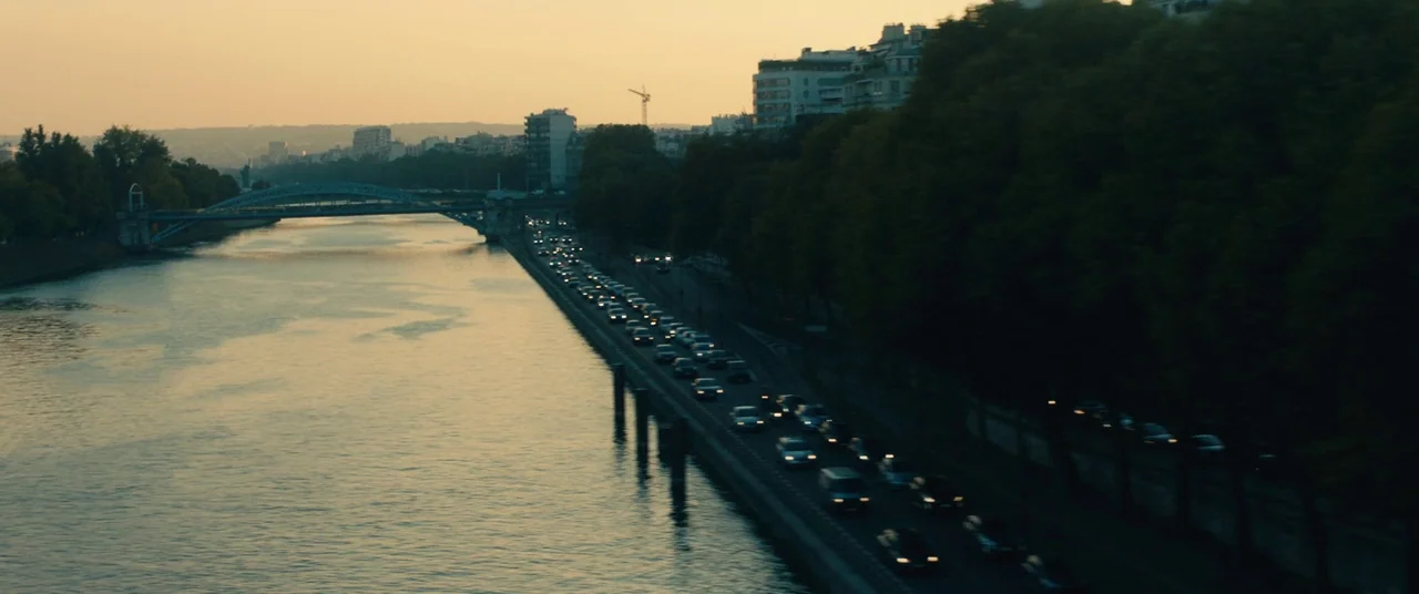 BPM (Beats Per Minute) (2017) movie still: establishing — A river flows alongside a busy urban highway during the golden hour in Paris. Rows of cars…