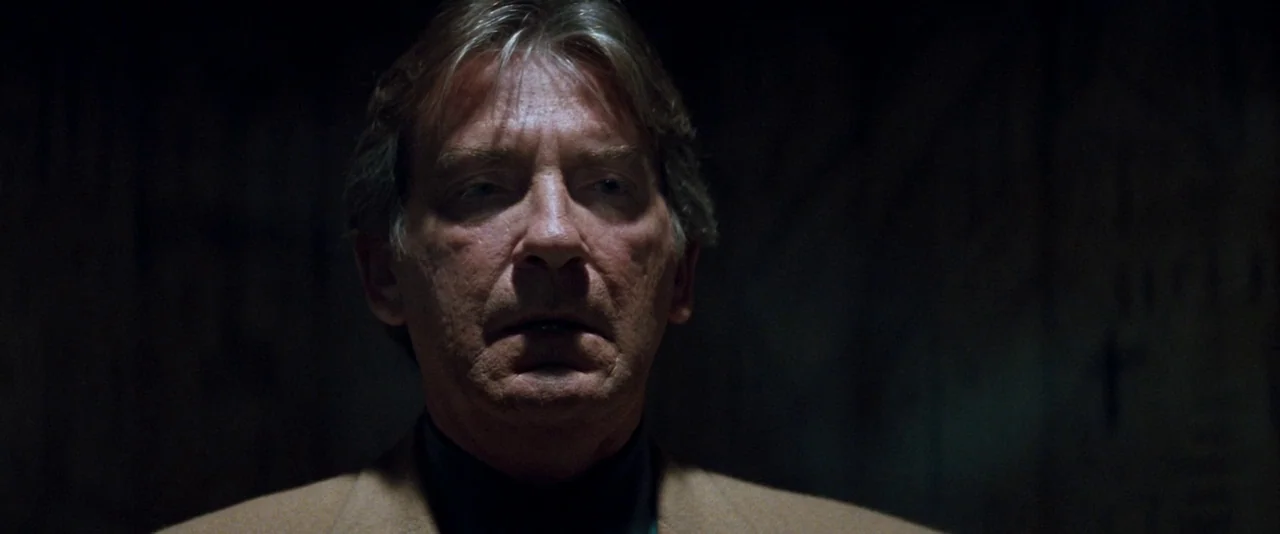 In The Mouth of Madness (1994) movie still: closeup — A man stands centered in a dark, indistinct interior environment. He faces the camera directly…