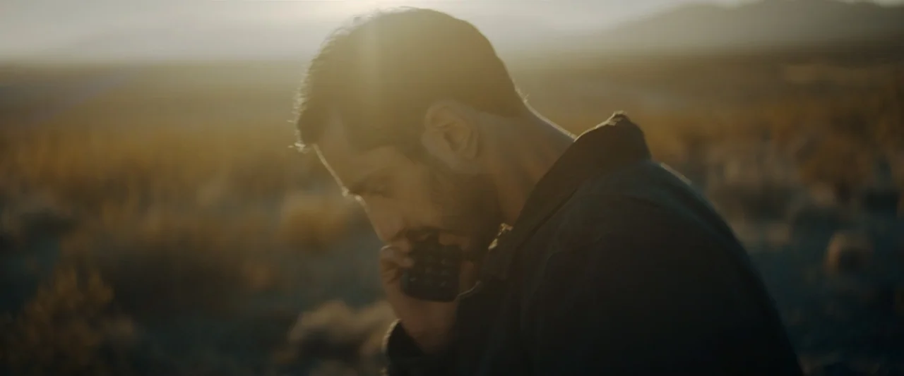 Encounter (2018) movie still: closeup — A man stands in an arid desert landscape during the late afternoon. He holds a black mobile phone to his ear…