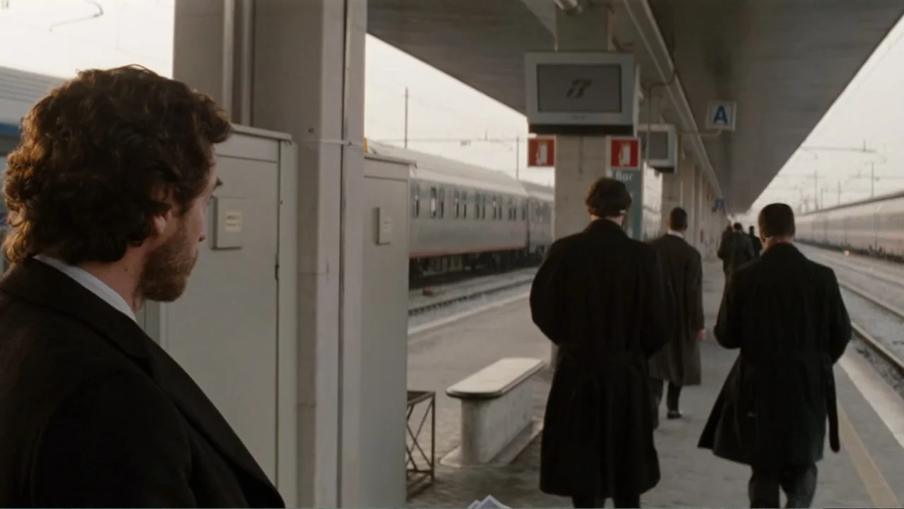 The Tourist (2010) movie still: medium — A modern outdoor train platform with concrete pillars and waiting areas. A man stands in the foreground…