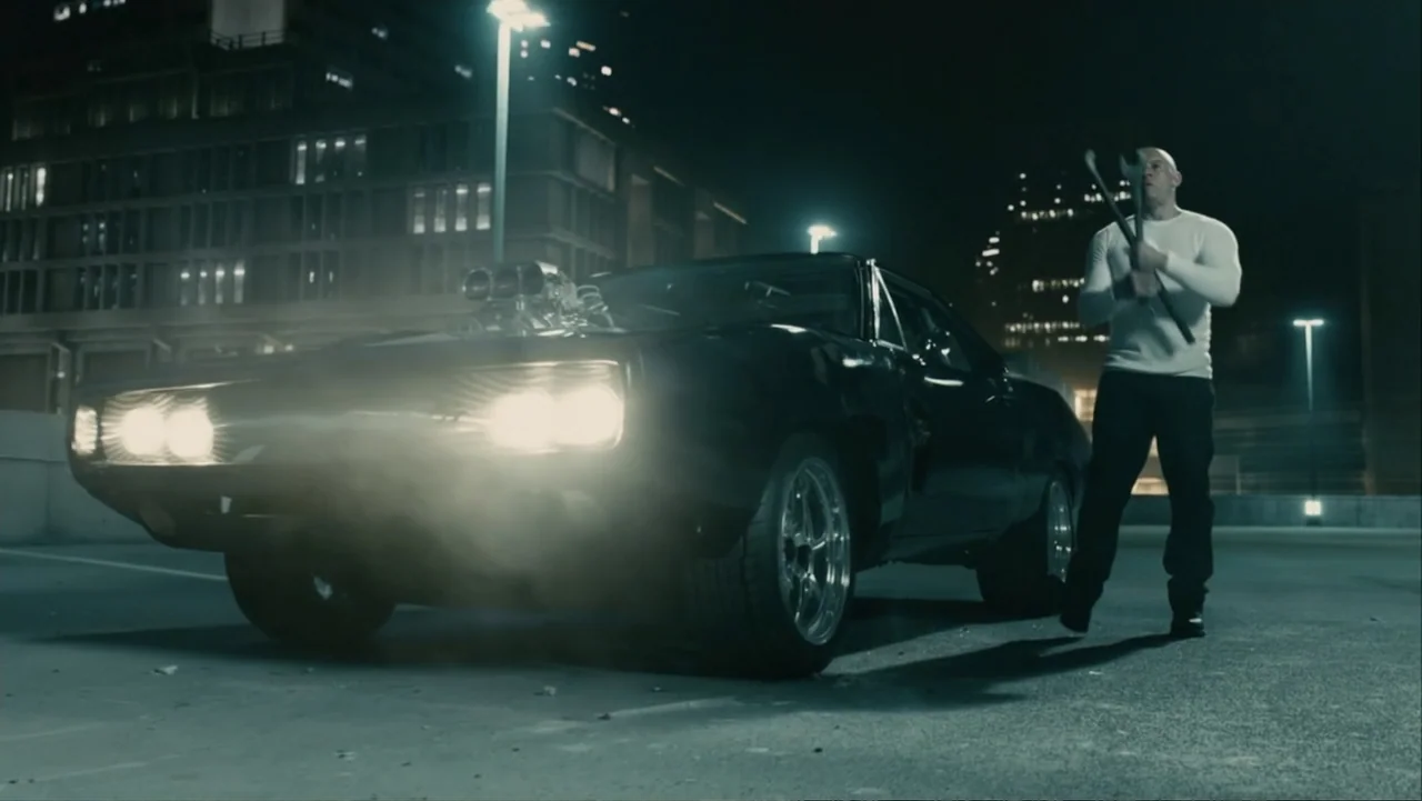 Furious 7 (2015) movie still: wide — A concrete parking garage level at night illuminated by powerful front-facing headlights. A bald man in a white…