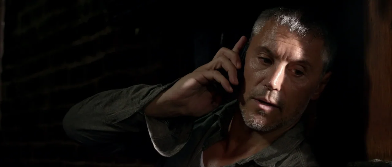 Rage (2014) movie still: closeup — A man stands in a dimly lit, shadowed room against a dark surface. The man holds a mobile phone to his ear during…
