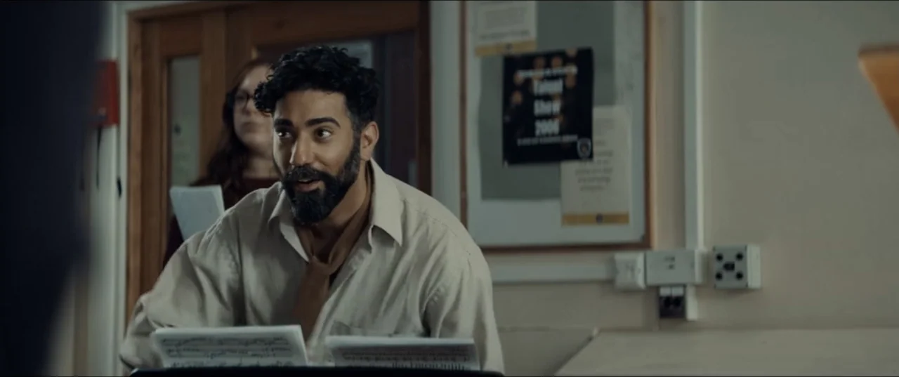 Bonus Track (2024) movie still: medium — A cluttered interior space with a cork notice board on the beige wall. A man sits at a music stand with…