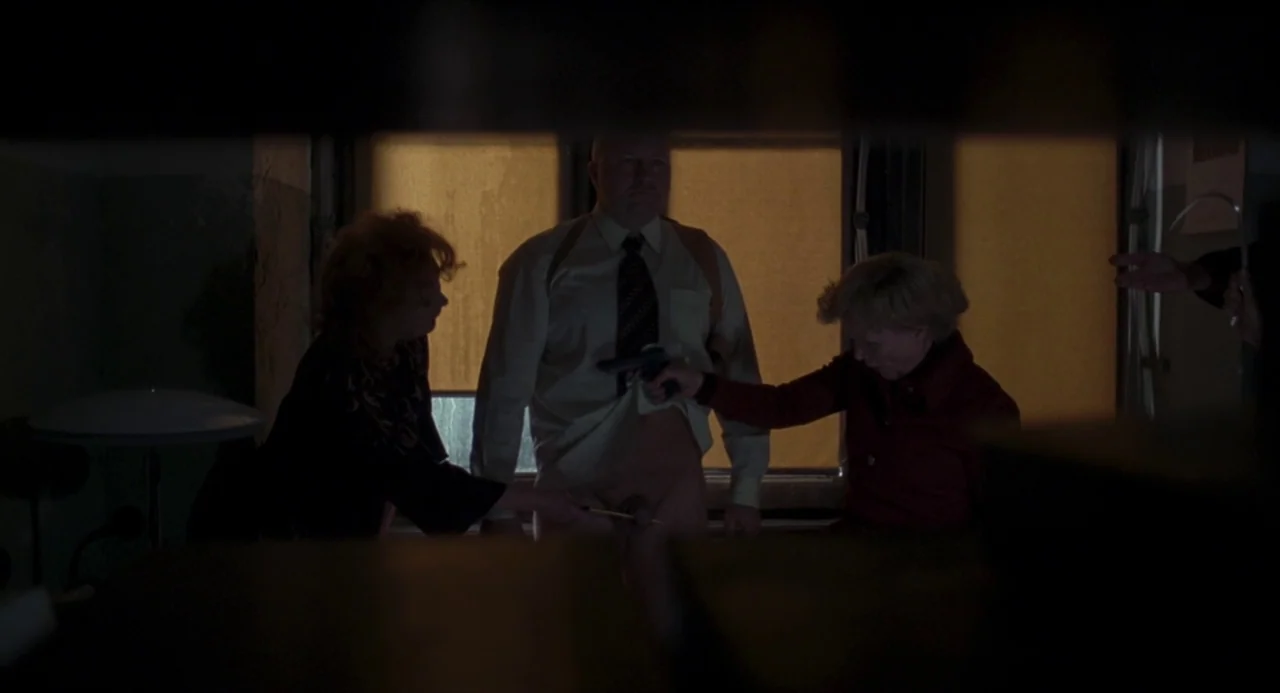Suspiria (2018) movie still: medium — A dimly lit room features a window covered by amber blinds in the background. A man stands between two women…