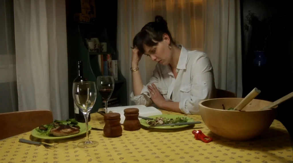 Humpday (2009) movie still: medium — A woman sits at a yellow table in a dimly lit home setting. She leans her head on her hand while reading an…