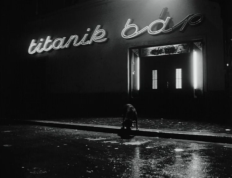 Damnation (1988) movie still: wide — A dark, wet asphalt surface fronts a building exterior at night. A glowing neon sign displays the name titanik…