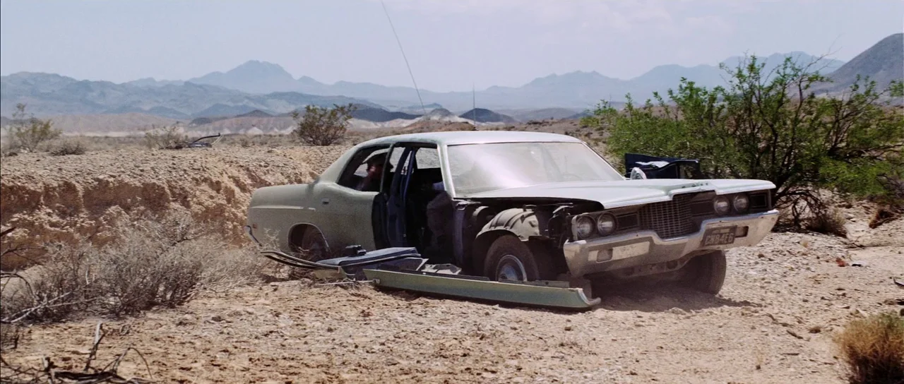 Diamonds Are Forever (1971) movie still: wide — An abandoned, damaged sedan rests on a rocky, scrub-filled arid terrain under clear skies. The car…