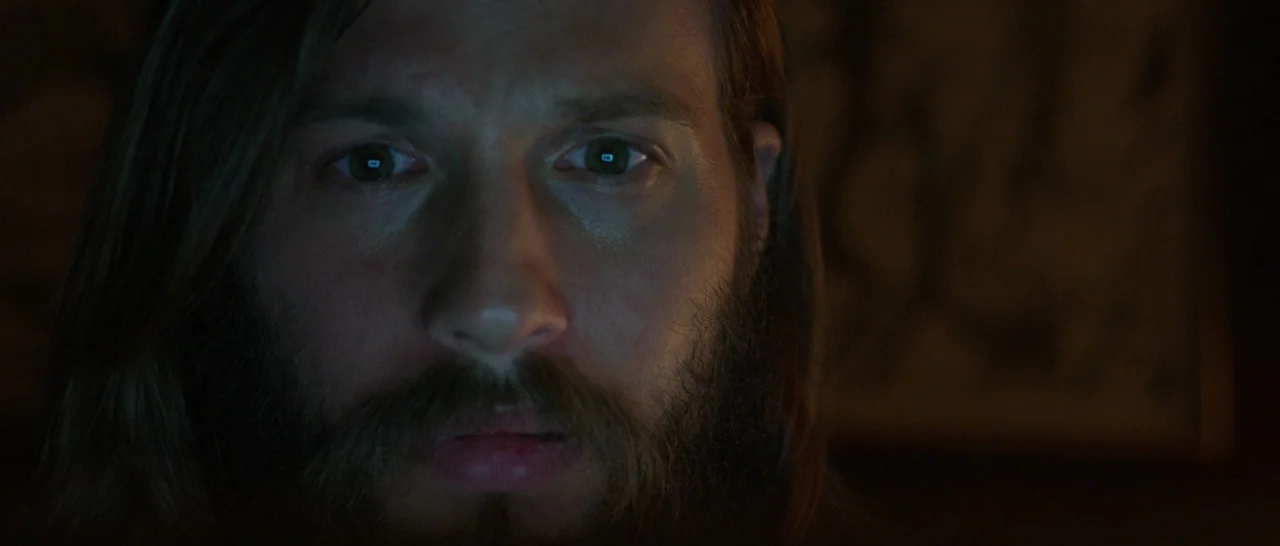 The Invitation (2015) movie still: closeup — A dimly lit interior space at night with a dark, indistinct background. A man with long hair and a…