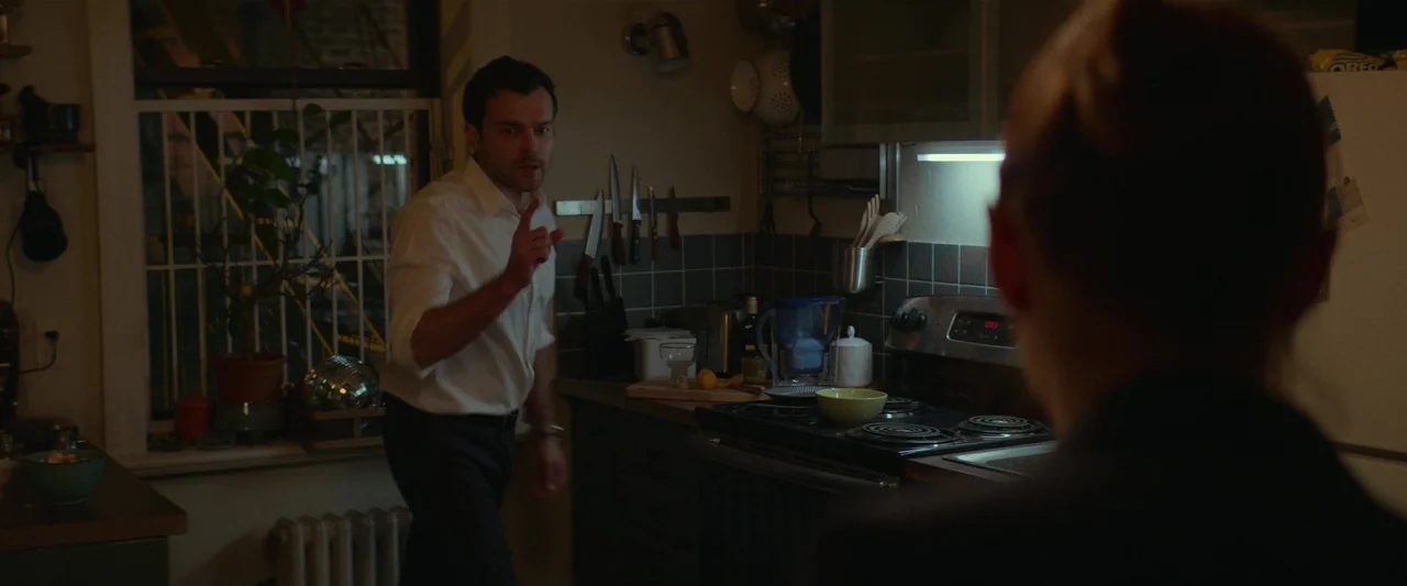 Fair Play (2023) movie still: medium — A cramped apartment area with a metal knife rack, stove, and a window view. A man in a white shirt stands…