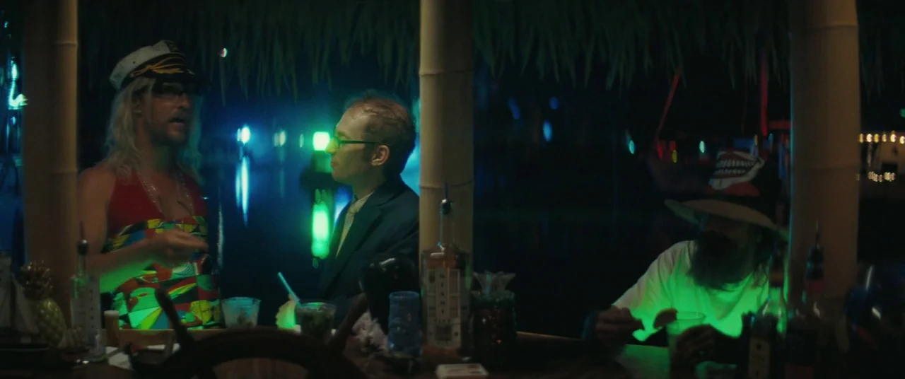 The Beach Bum (2019) movie still: wide — A dark tiki bar at night features bamboo supports and colorful neon accent lighting. Two men in foreground…