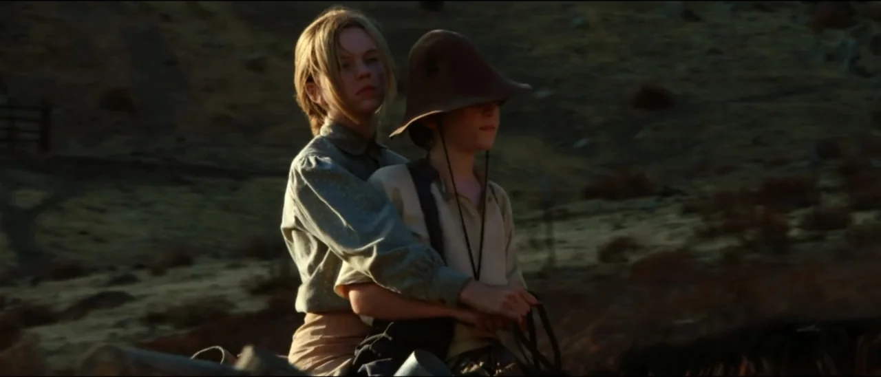 Dead for a Dollar (2022) movie still: medium — An open arid landscape with sparse dry brush during the golden hour light. A young woman holds a…