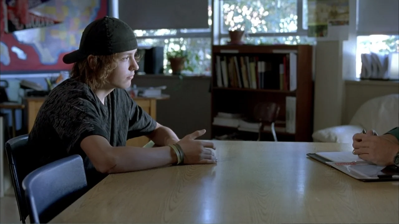 Paranoid Park (2007) movie still: medium — An office interior features a wooden table and a cluttered bookshelf in the background. A young man sits…