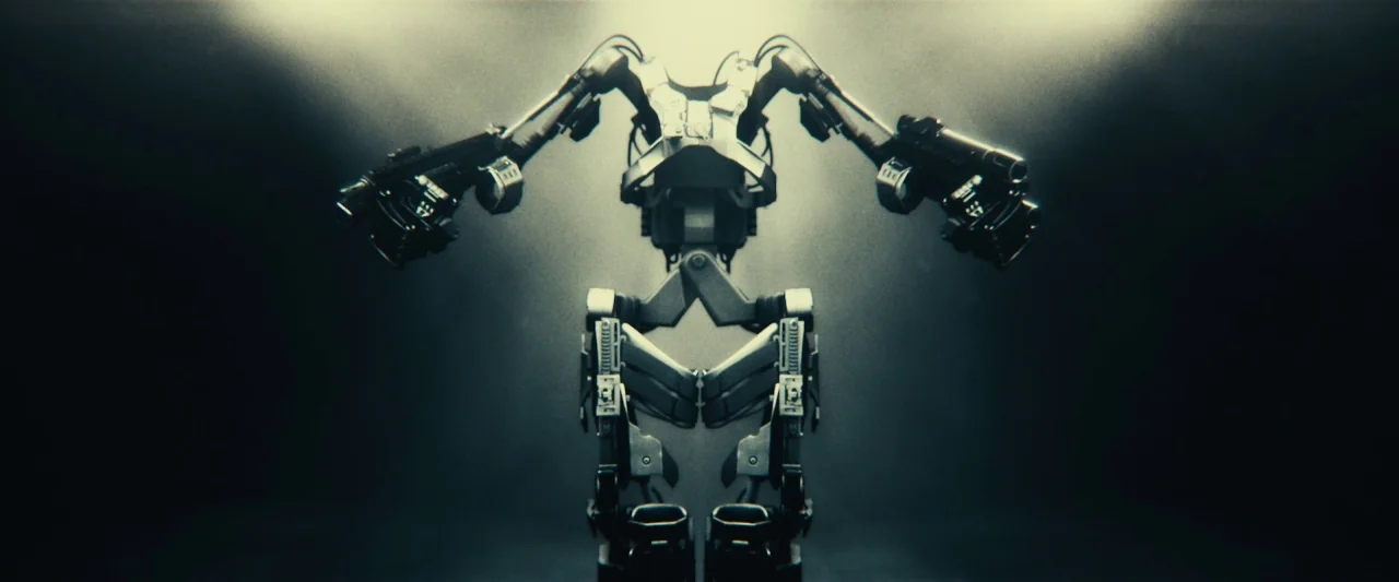 Edge of Tomorrow (2014) movie still: medium — A complex metallic mechanical exoskeleton stands in a dark, minimalist industrial environment. The…