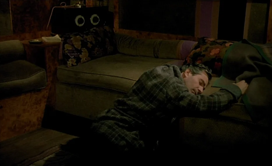 Mélo (1986) movie still: medium — A small, dimly lit interior space with wood-paneled walls and a fabric-covered sofa. A man in a plaid jacket rests…